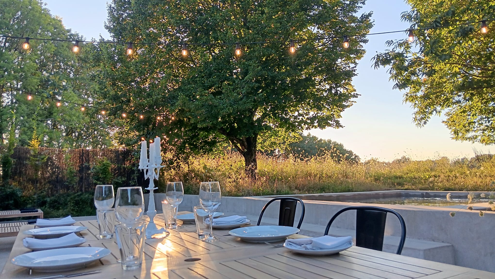 La Maison Nid, terrace, evening setting, candles lit on set outdoor table, pool, trees and field behind.